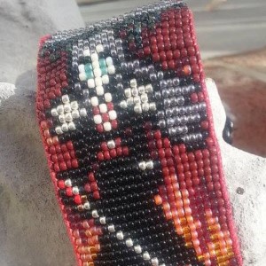 A bracelet of beadwork, depicts a man in black with a long sword standing in front of flames.